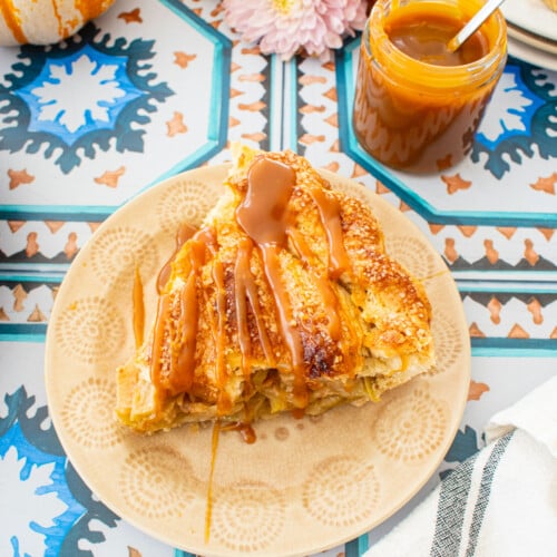 slice of caramel apple pie with a drizzle of caramel sauce on top on a tile table with mini pumpkins and fresh flowers.