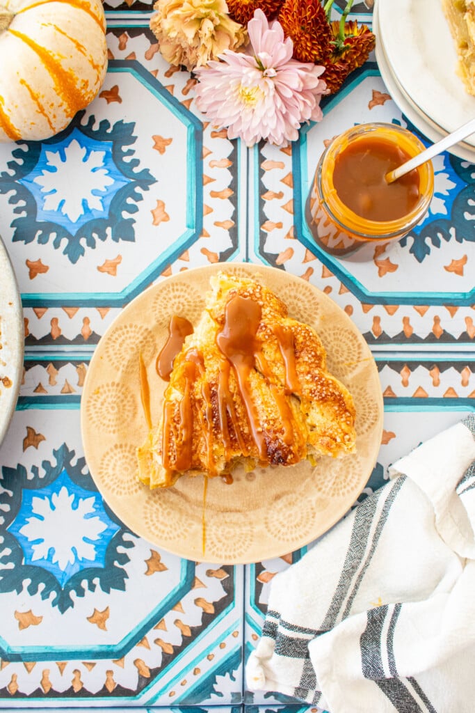 slice of caramel apple pie on a dessert plate with a jar of caramel, mini pumpkins and fresh flowers on a tile table.