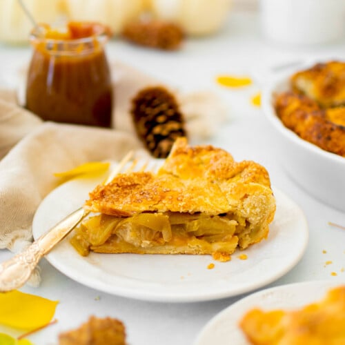 slice of caramel apple pie on a plate on a white counter with fall leaves, pinecones and white pumpkins.