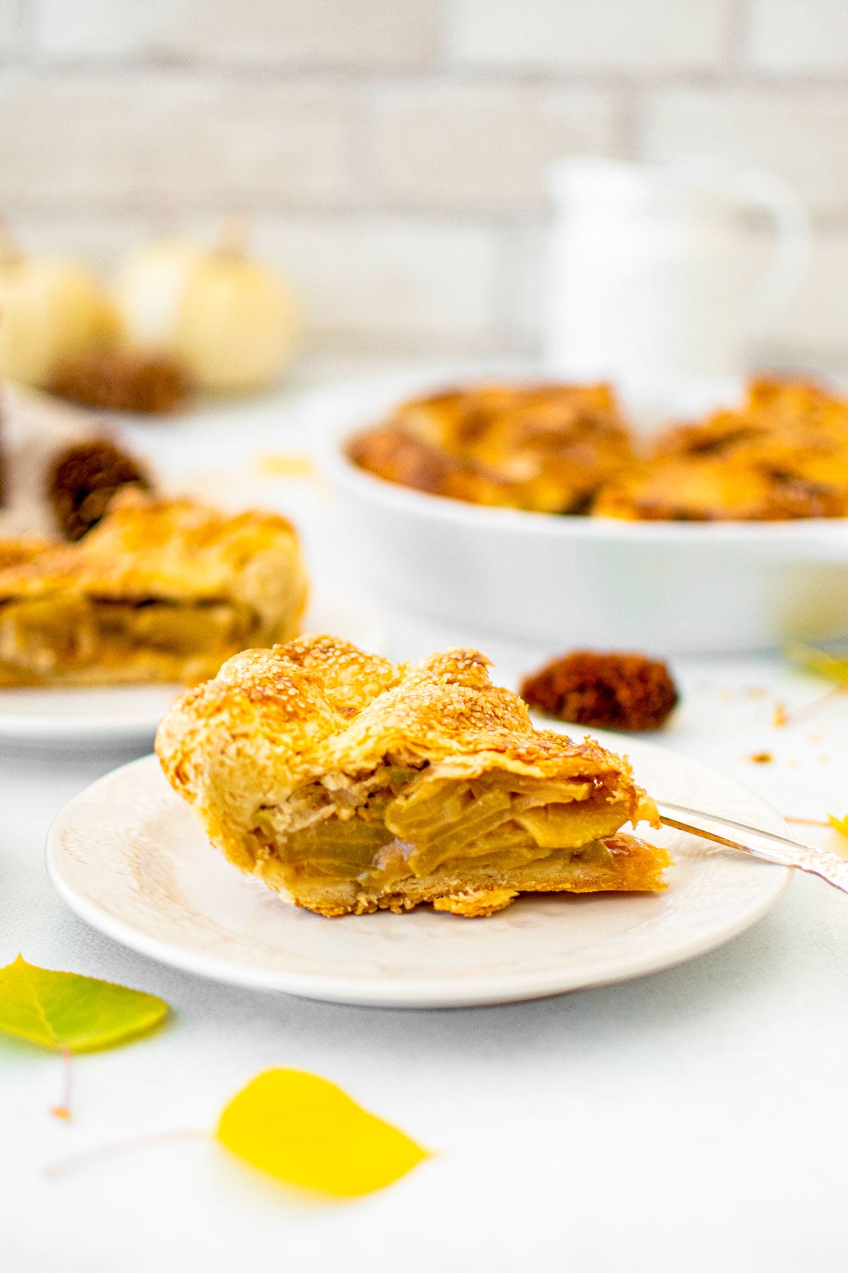 slice of caramel apple pie on a plate on a white counter with fall leaves, pinecones and white pumpkins.