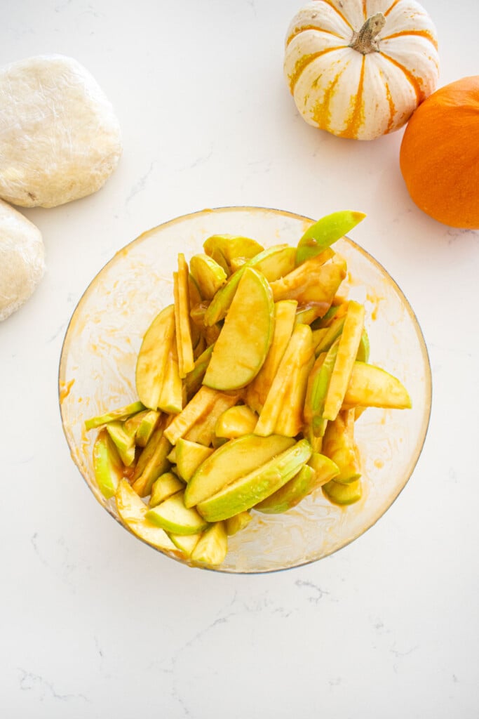 glass bowl of apples tossed with caramel sauce on a marble counter with mini pumpkins and pie crust.