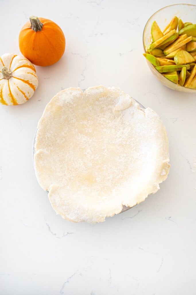 pie pan with pie crust placed inside sitting on a marble counter with a bowl of apple pie filling and mini pumpkins.