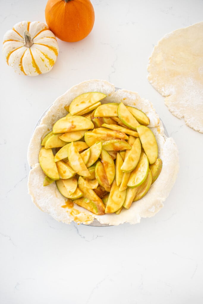 pie crust in a pie pan with caramel apple filling spread evenly sitting on a marble counter with mini pumpkins.