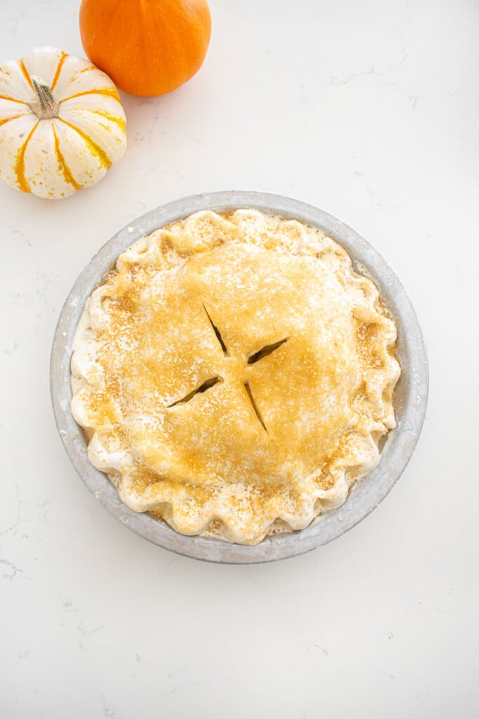 caramel apple pie brushed with heavy whipping cream and sprinkled with turbinado sugar ready to be baked sitting on a marble counter with mini pumpkins.