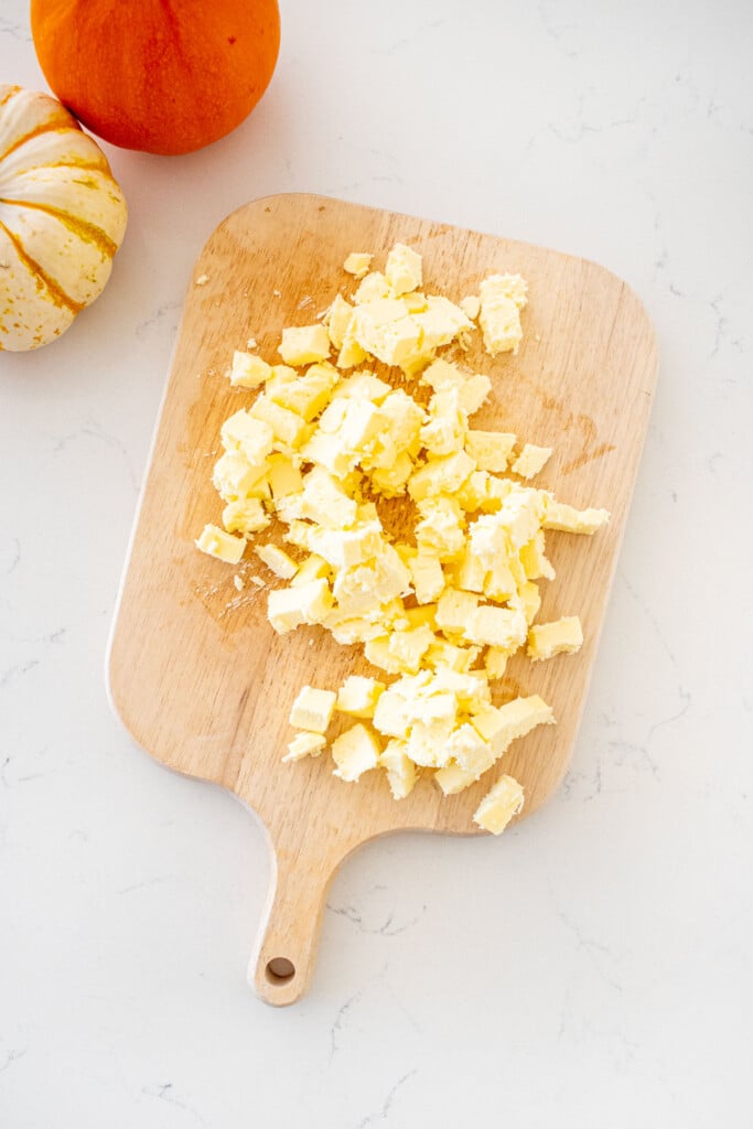 wood cutting board with cold butter chopped into cubes on a marble counter with mini pumpkins.