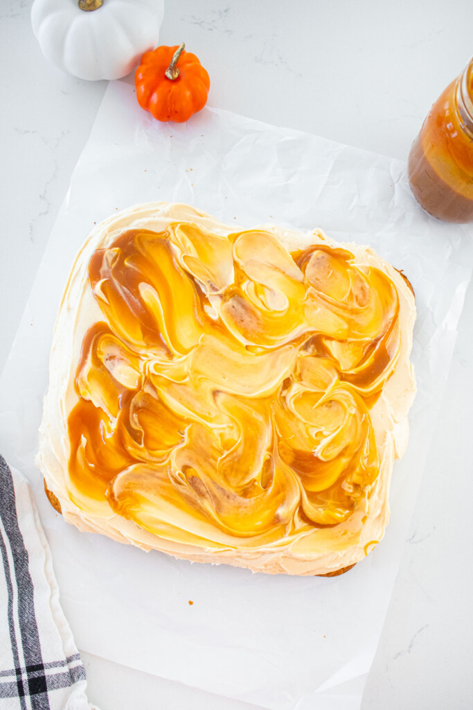 square pumpkin spice snack cake on a piece of parchment paper on a marble counter with caramel frosting swirled around.