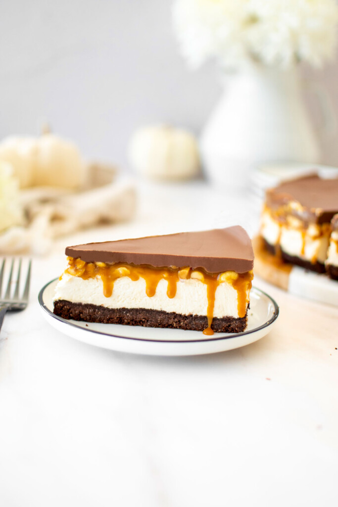 eggless snickers cheesecake slice on a plate on a marble counter with white pumpkins.