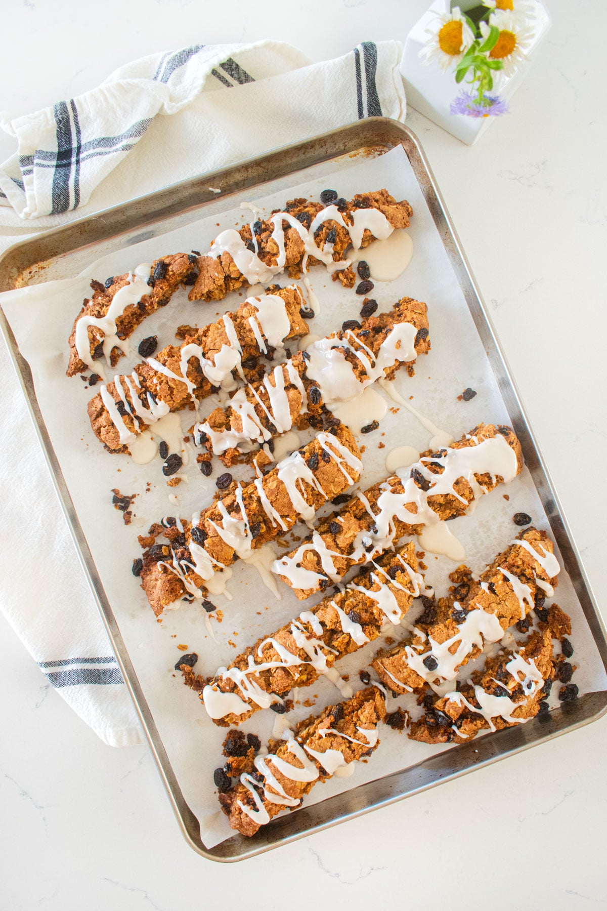 glazed oatmeal raisin biscotti on a baking sheet on a marble counter with a small vase of flowers.