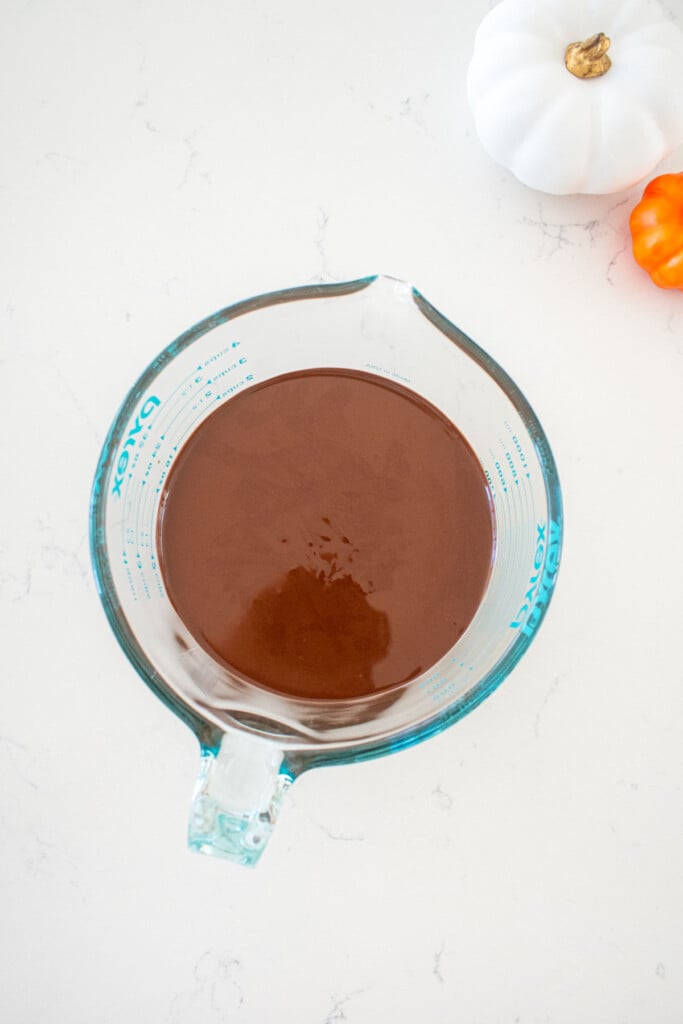 glass liquid measuring cup with melted milk chocolate on a marble counter with small pumpkins.
