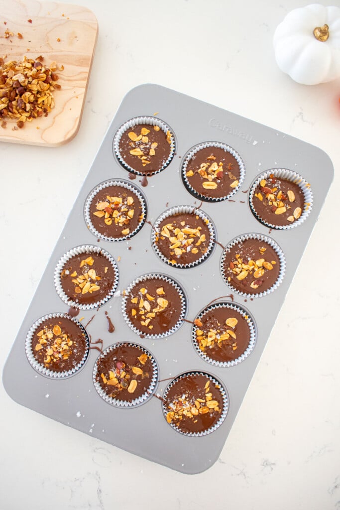cupcake pan with milk chocolate filled to the top with chopped peanuts and flaky sea salt on top on a marble counter.