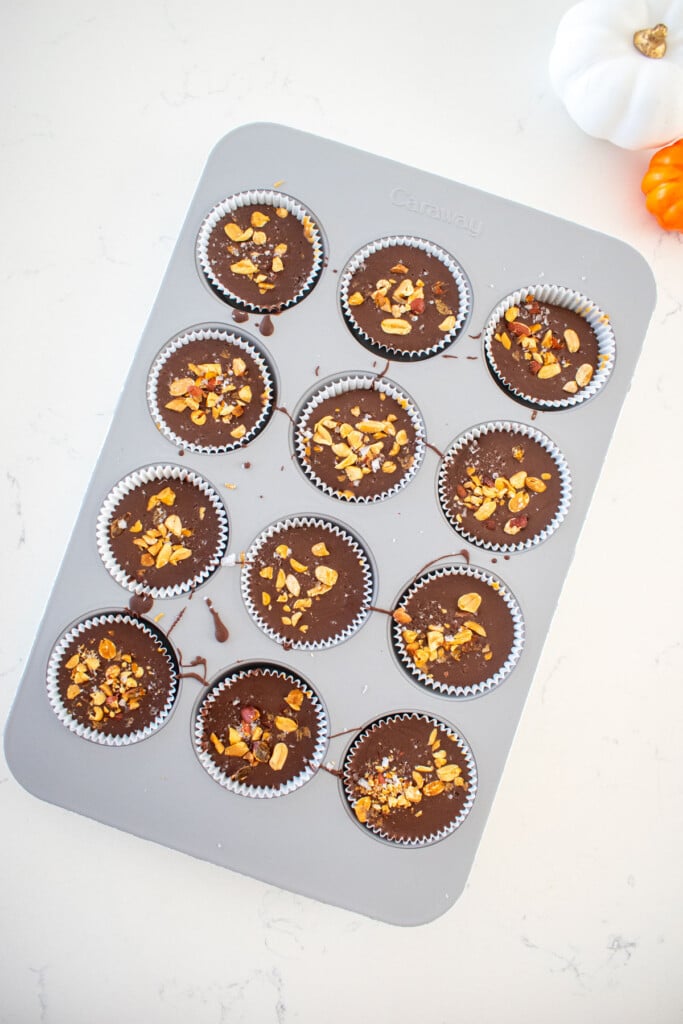 cupcake pan with milk chocolate filled to the top with chopped peanuts and flaky sea salt on top on a marble counter.