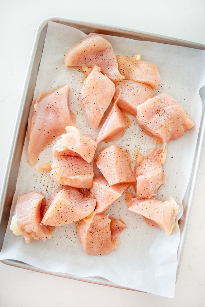 raw chicken breasts cut into cubes on a baking sheet on a white marble counter.