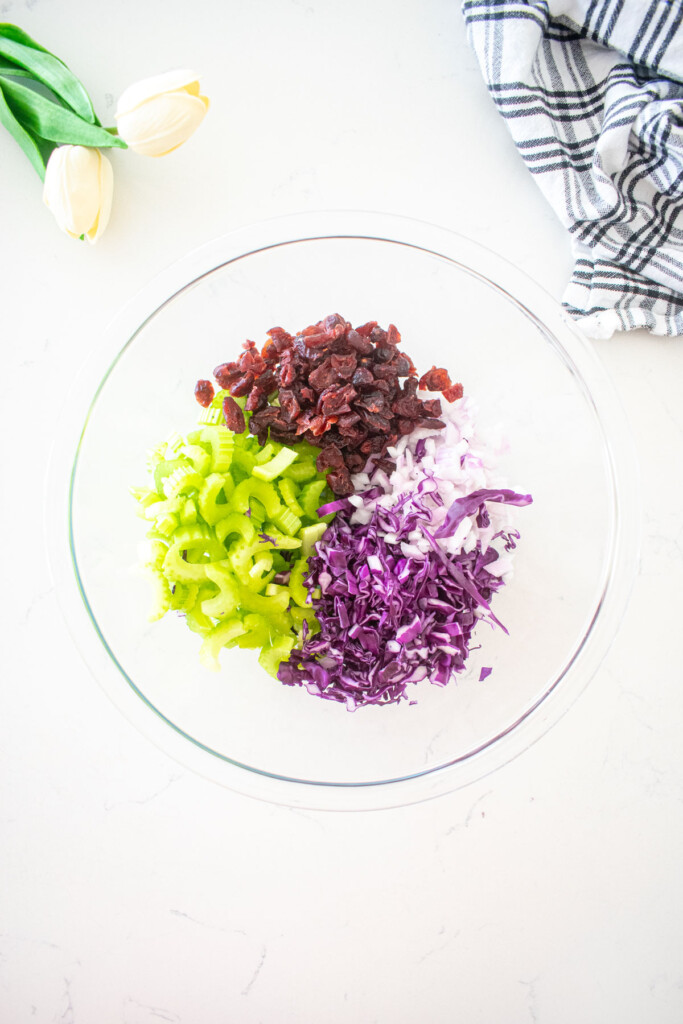 chopped celery, cabbage, red onion and dried cranberries in a glass mixing bowl on a white marble counter.