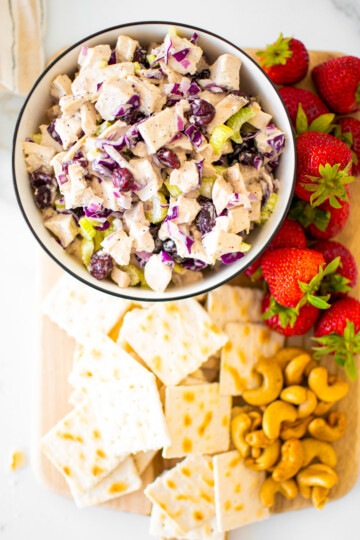 healthy honey chicken salad in a bowl on a board with crackers, nuts and strawberries.