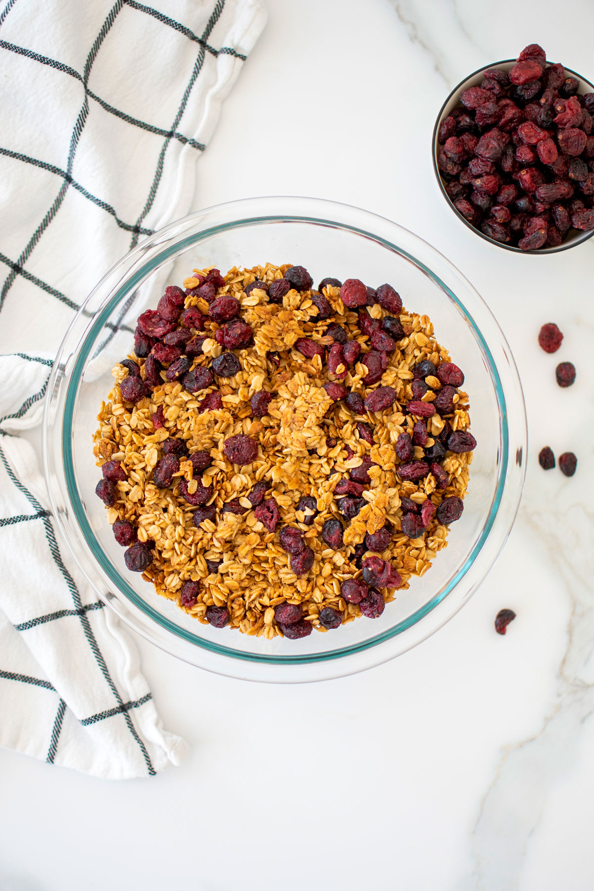 homemade vanilla granola with cranberries in a glass mixing bowl on a white marble counter.