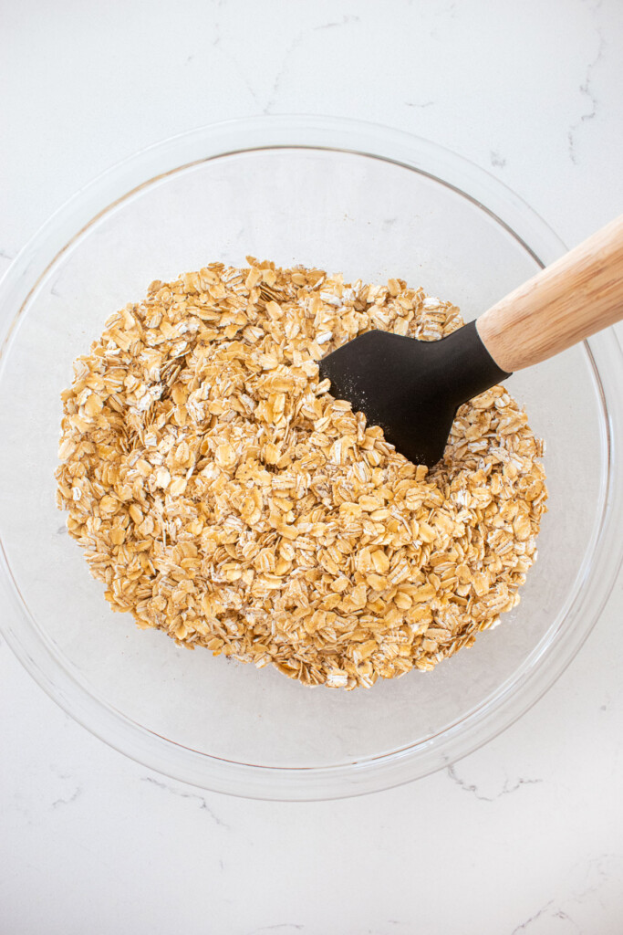 dry ingredients for homemade vanilla granola in a glass mixing bowl with a spatula on a white marble counter.