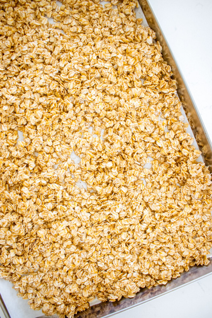 homemade vanilla granola spread out on a baking sheet on a white marble counter.