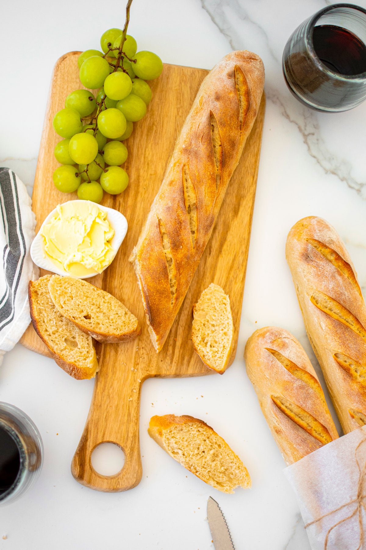 organic french baguette on a wood serving board with butter and grapes on a marble table with wine glass.