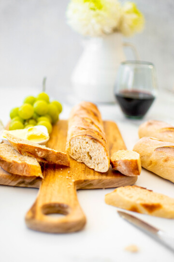 organic french baguette on a wood serving board with butter and grapes on a marble table with wine glass.