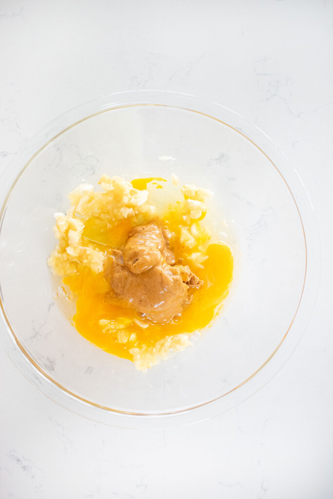 banana, egg, and peanut butter in a glass mixing bowl on a white marble countertop.
