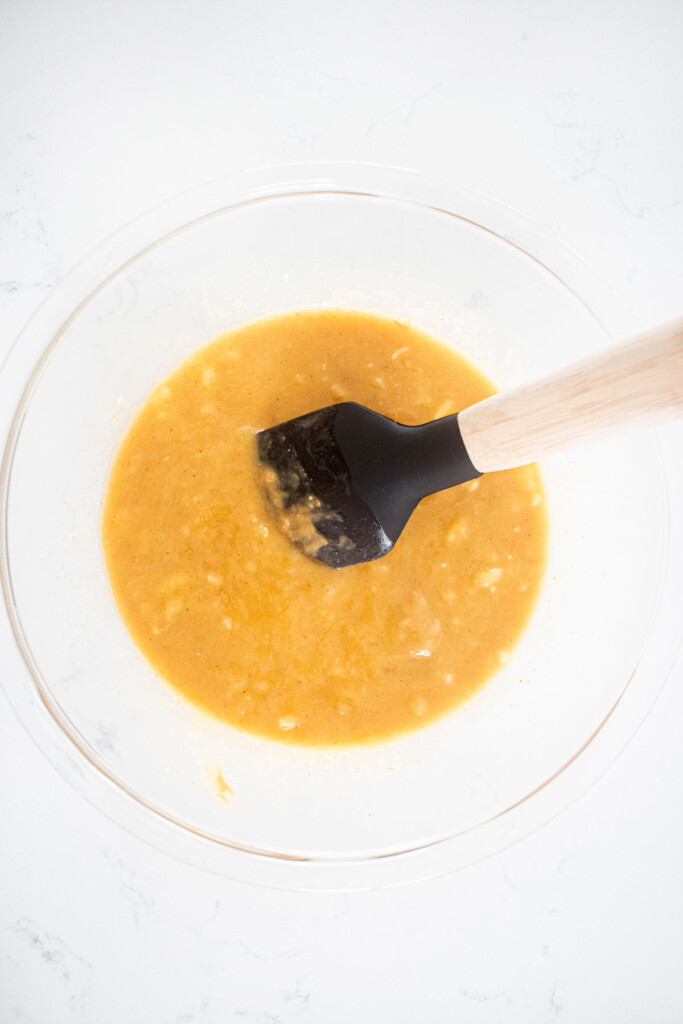 peanut butter banana dog treats being mixed together in a glass mixing bowl with a spatula on a white countertop.