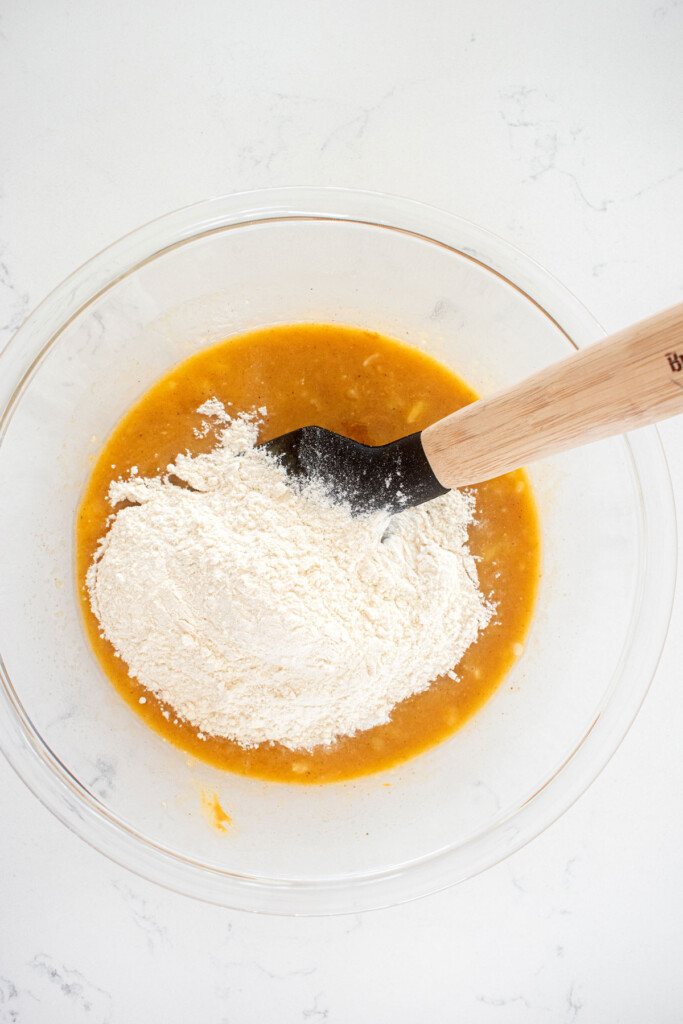 peanut butter banana dog treats being mixed together in a glass mixing bowl with a spatula on a white countertop.