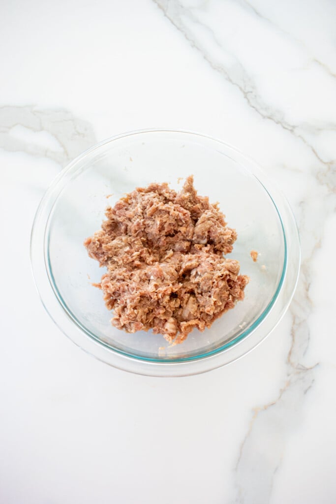 ground pork with with spices mixed in a glass mixing bowl on a white marble counter.