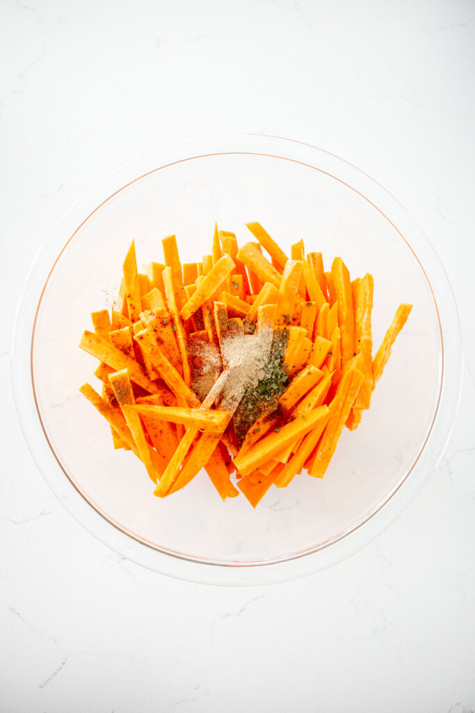 sweet potato fries with spices in a glass mixing bowl on a white marble countertop.