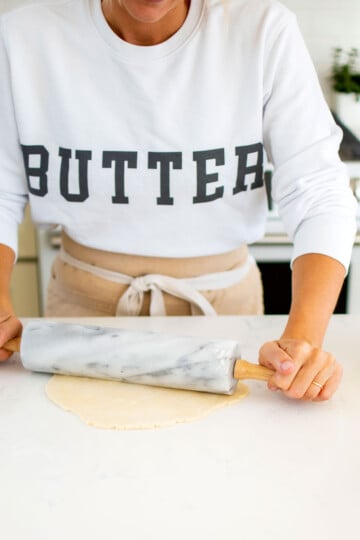 woman wearing a butter sweatshirt rolling out a pie crust on a white kitchen counter.