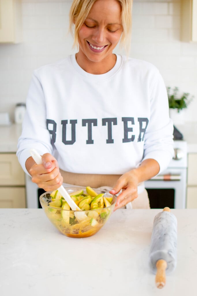 woman wearing a butter sweatshirt stirring a caramel apple filling in a glass mixing bowl at the kitchen counter.