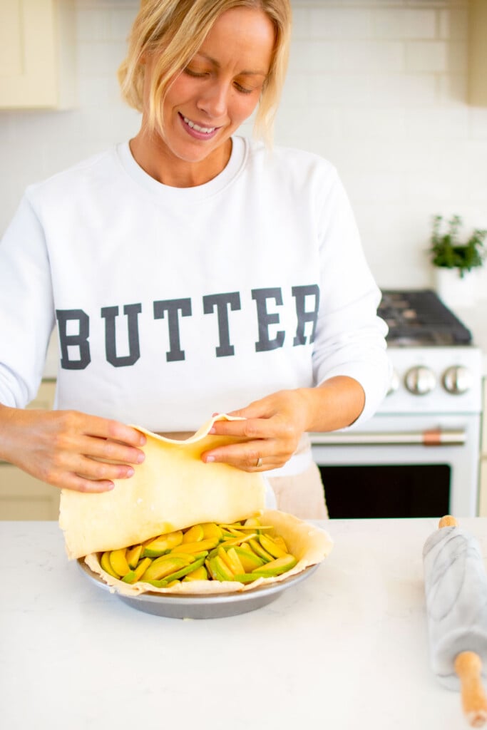 woman wearing a butter sweatshirt adding a top crust onto a caramel apple pie at kitchen counter.