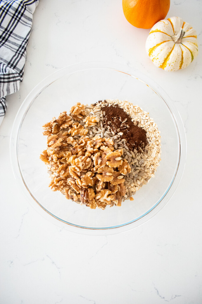 dry ingredients for vanilla granola in a glass mixing bowl on a marble counter with tiny pumpkins and a kitchen towel.