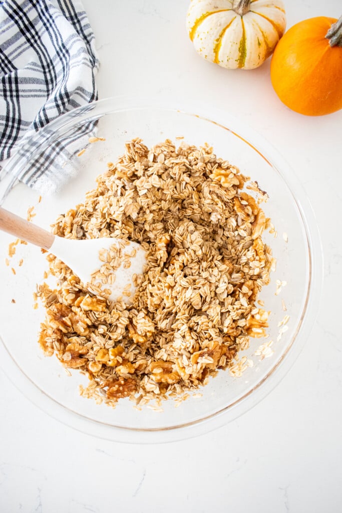 glass mixing bowl with vanilla granola mixed together with a spatula on a marble counter.