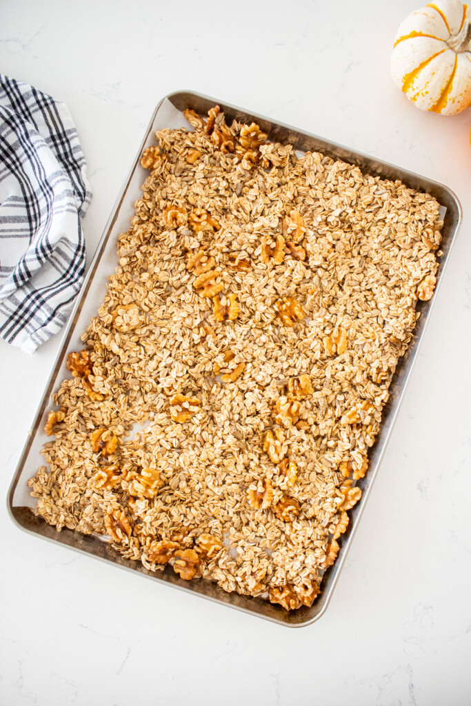 baking sheet with vanilla granola spread out evenly on a marble counter with a kitchen towel and tiny pumpkin.