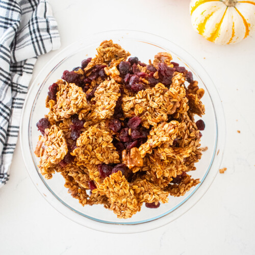 big glass bowl filled with big clusters of vanilla granola with cranberries on a marble counter with tiny pumpkins and a kitchen towel.