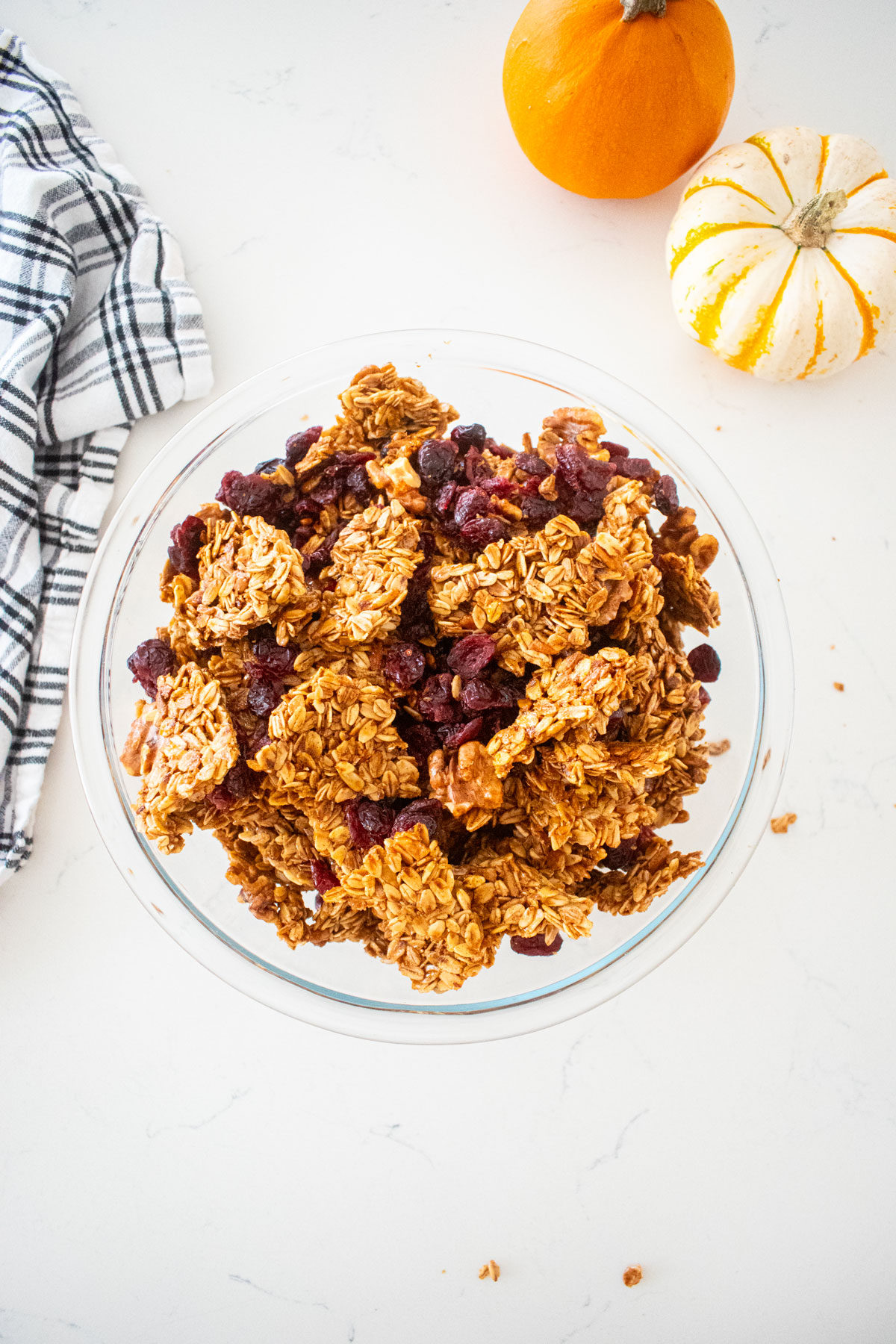 big glass bowl filled with big clusters of vanilla granola with cranberries on a marble counter with tiny pumpkins and a kitchen towel.