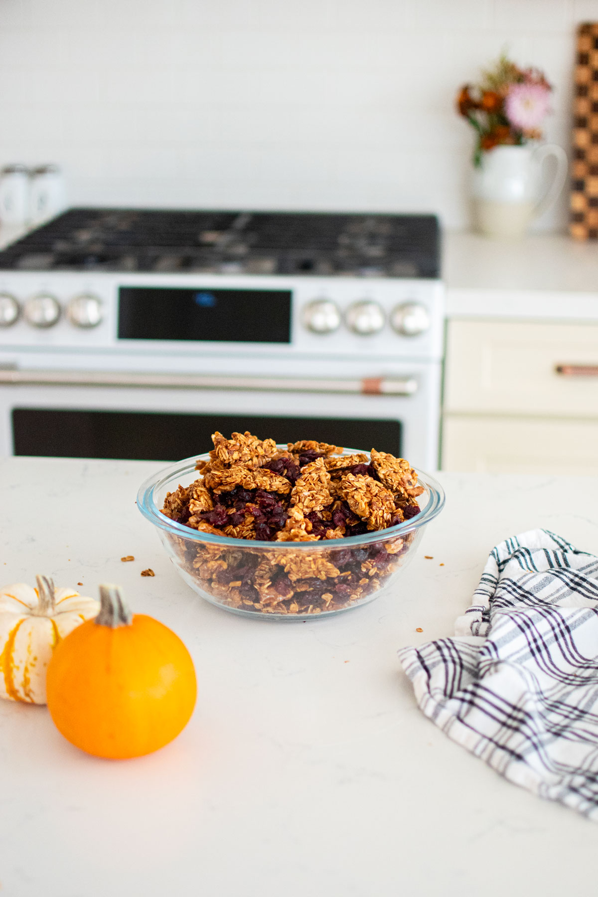 glass bowl of homemade vanilla granola with cranberries on a white marble counter with tiny pumpkins and a kitchen towel.