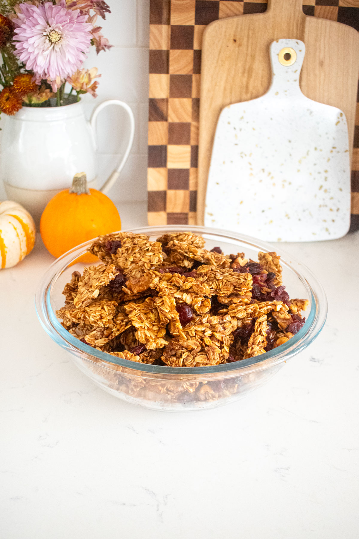 glass bowl of vanilla granola with cranberries on a marble kitchen counter with tiny pumpkins and a vase of flowers.