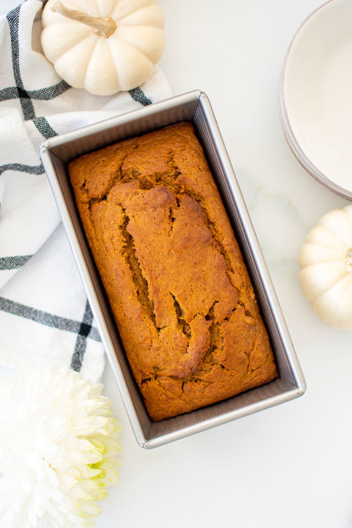 vegan pumpkin spice banana bread in an aluminum loaf pan on a white marble counter with white pumpkins and flowers.