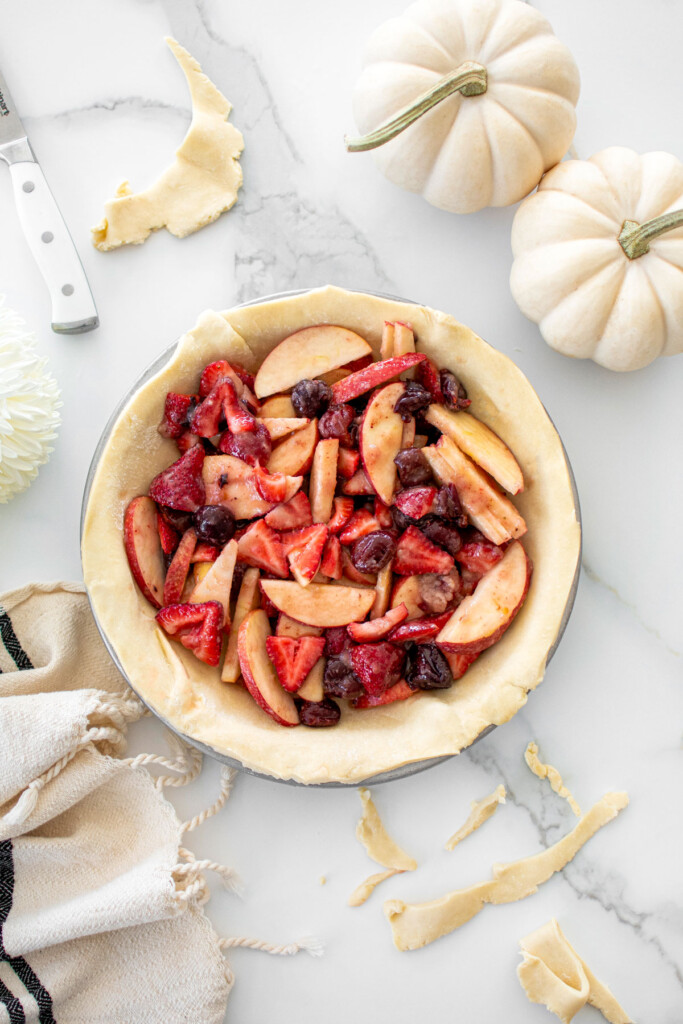 apple berry pie being filled on a white marble counter with white pumpkins and a dish towel.
