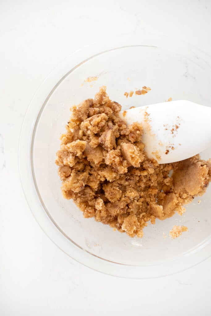 streusel topping mixed together in a glass mixing bowl with a spatula on a white marble counter.