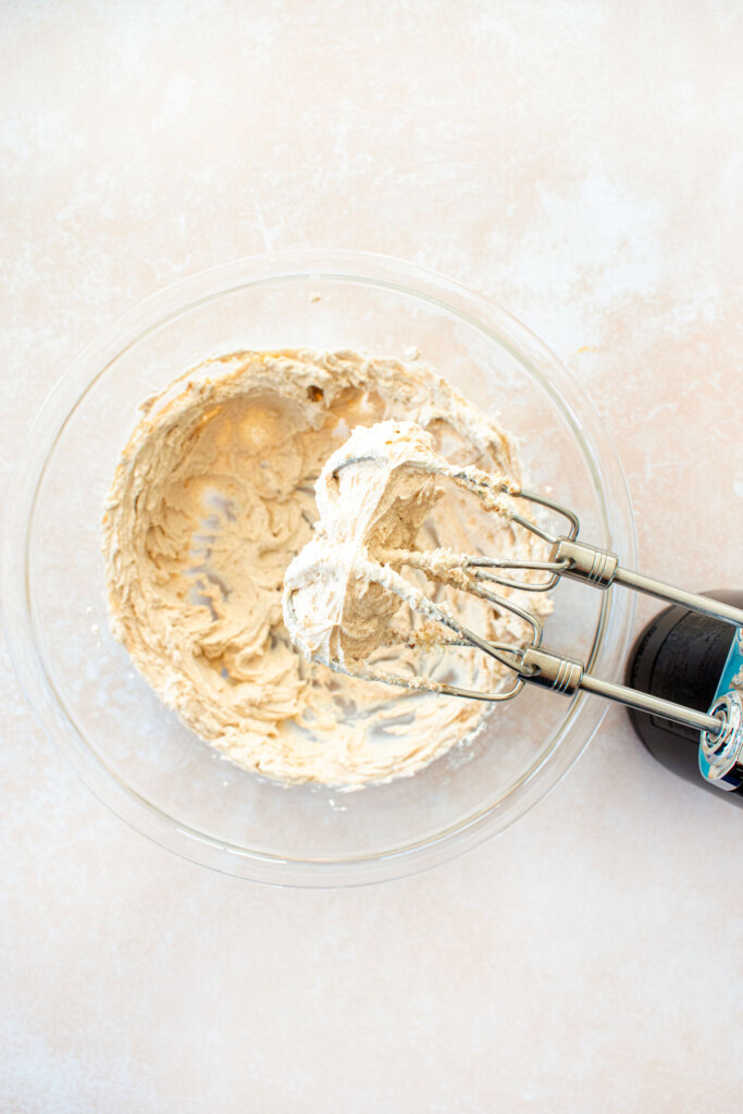 cinnamon honey cream cheese spread being mixed with a hand mixer in a glass bowl on a tan countertop.