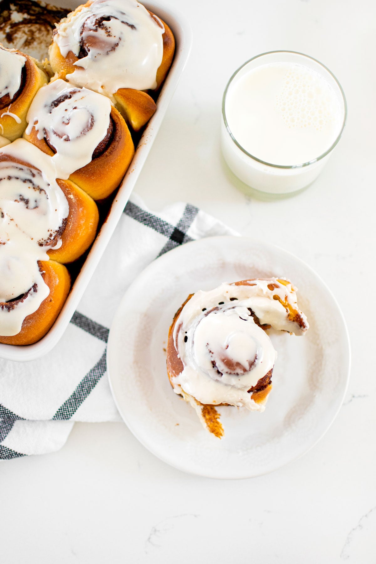 cinnamon roll on a white plate with a glass of milk on a white marble counter.