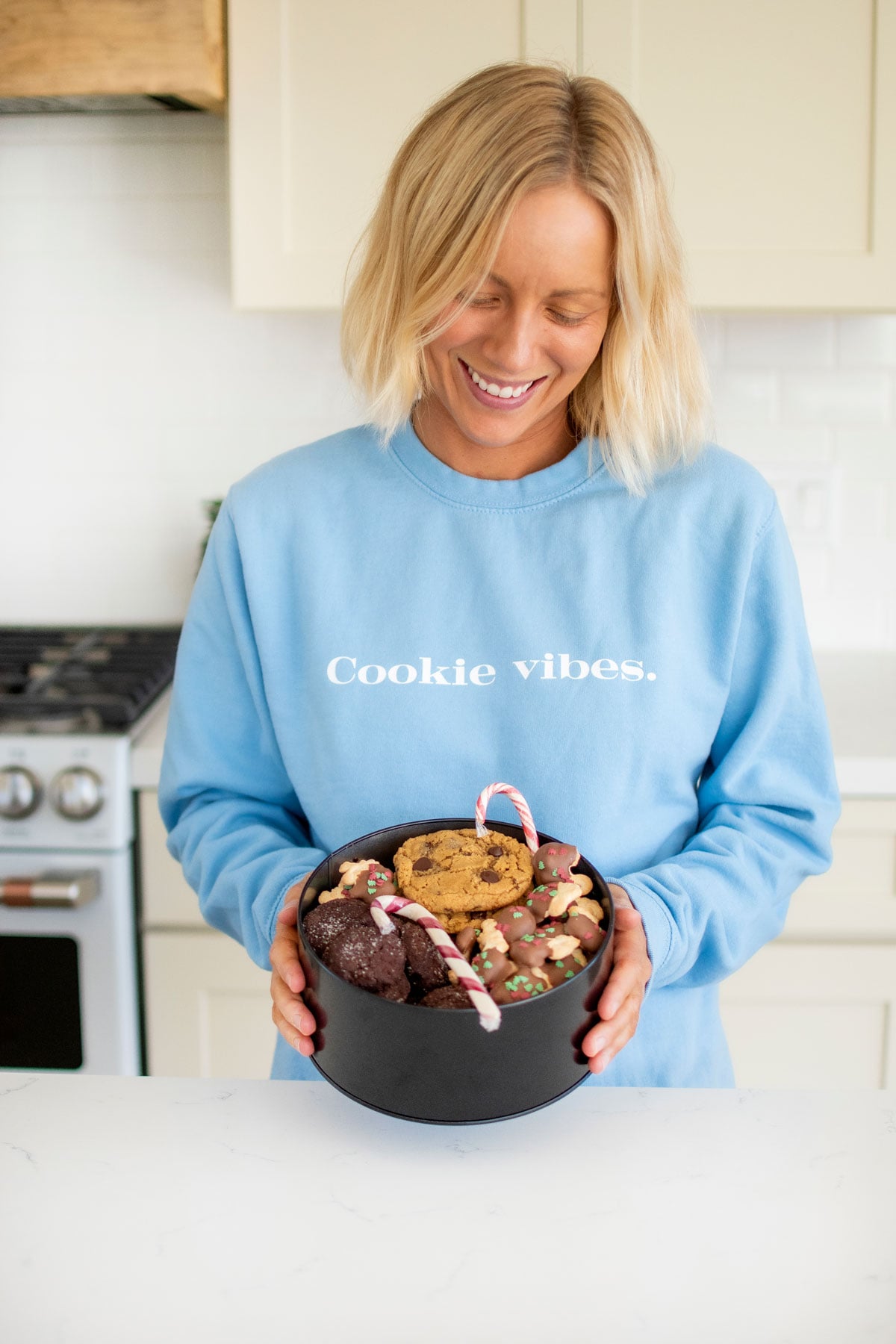 woman holding a tin of christmas cookies in the kitchen wearing a blue sweatshirt with cookie vibes on it.