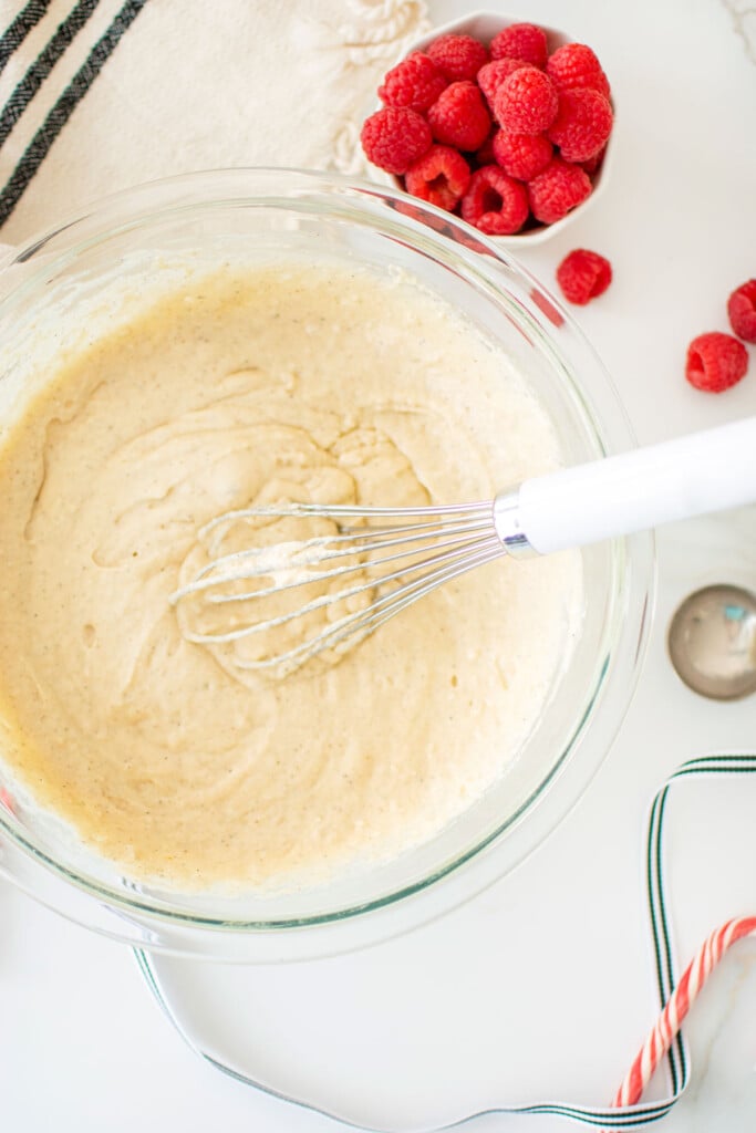 homemade pancake batter in a glass mixing bowl with a whisk on a marble counter with fresh raspberries.