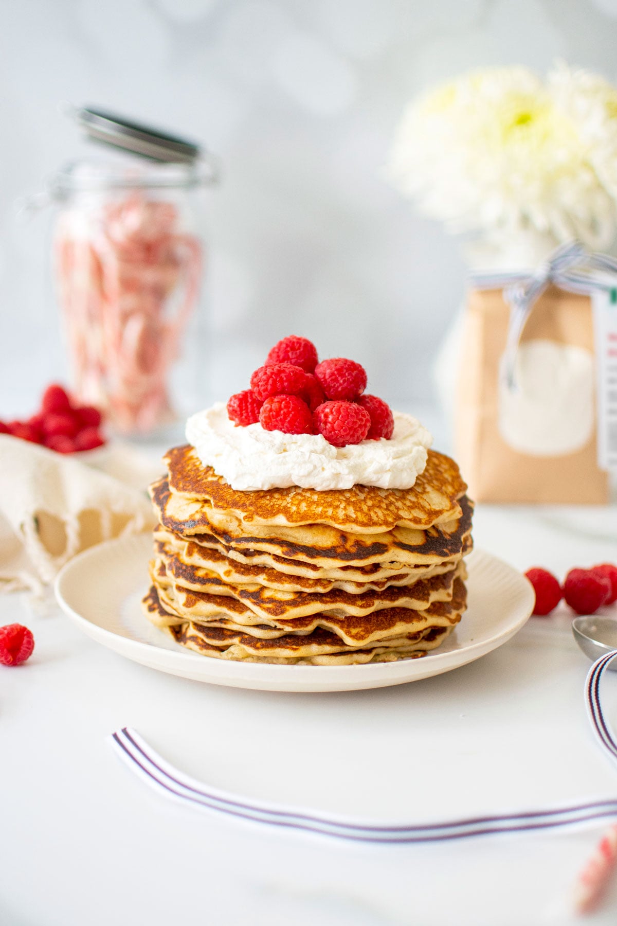stack of homemade pancakes with whipped cream and raspberries on a plate on a white marble counter.