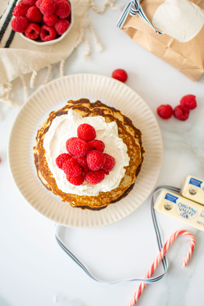 homemade pancakes with whipped cream and raspberries on a plate on a white marble counter.