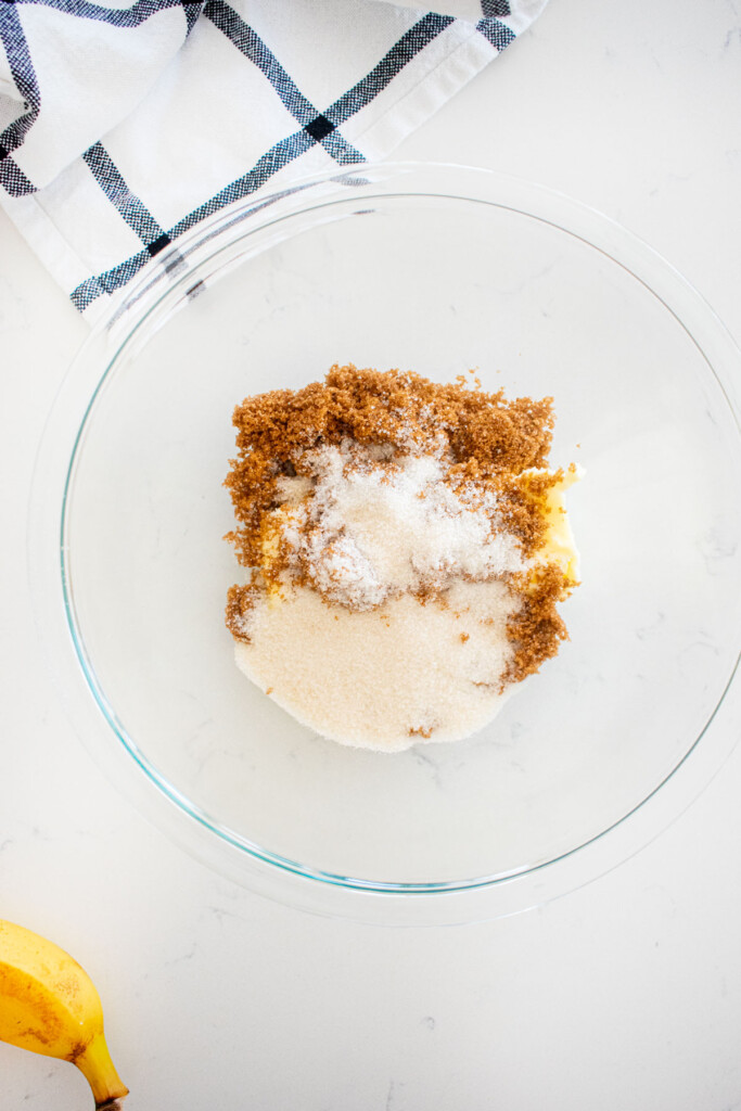 glass mixing bowl with butter, cane sugar and dark brown sugar on a white marble counter.