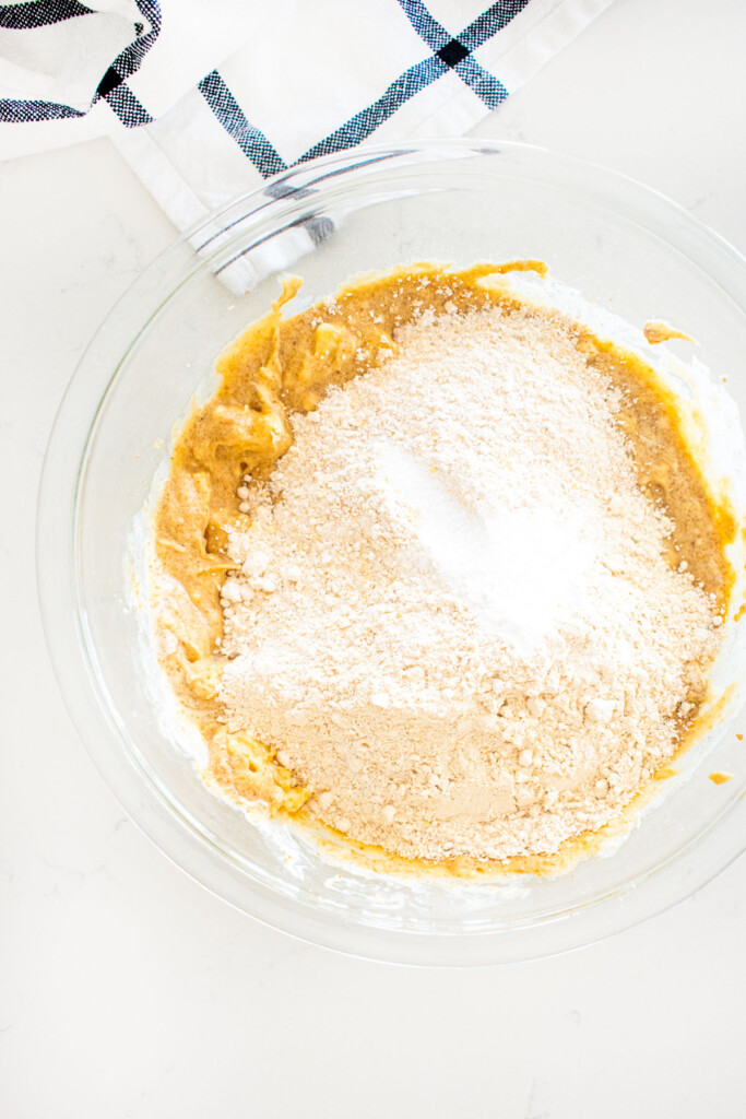 dry ingredients being added to wet ingredients for eggless banana bread in a glass mixing bowl on a marble counter.