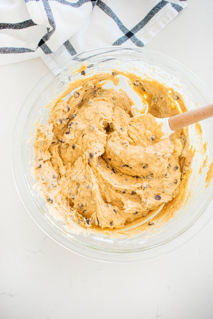 eggless mini chocolate chip banana bread batter with a spatula in a glass mixing bowl on a marble counter.